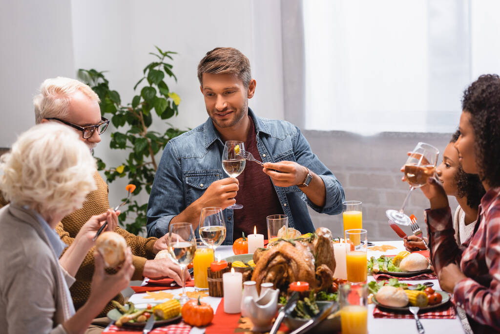 Enfoque selectivo del hombre sosteniendo el vino cerca de la familia multiétnica durante la cena de acción de gracias en casa  - Foto, imagen