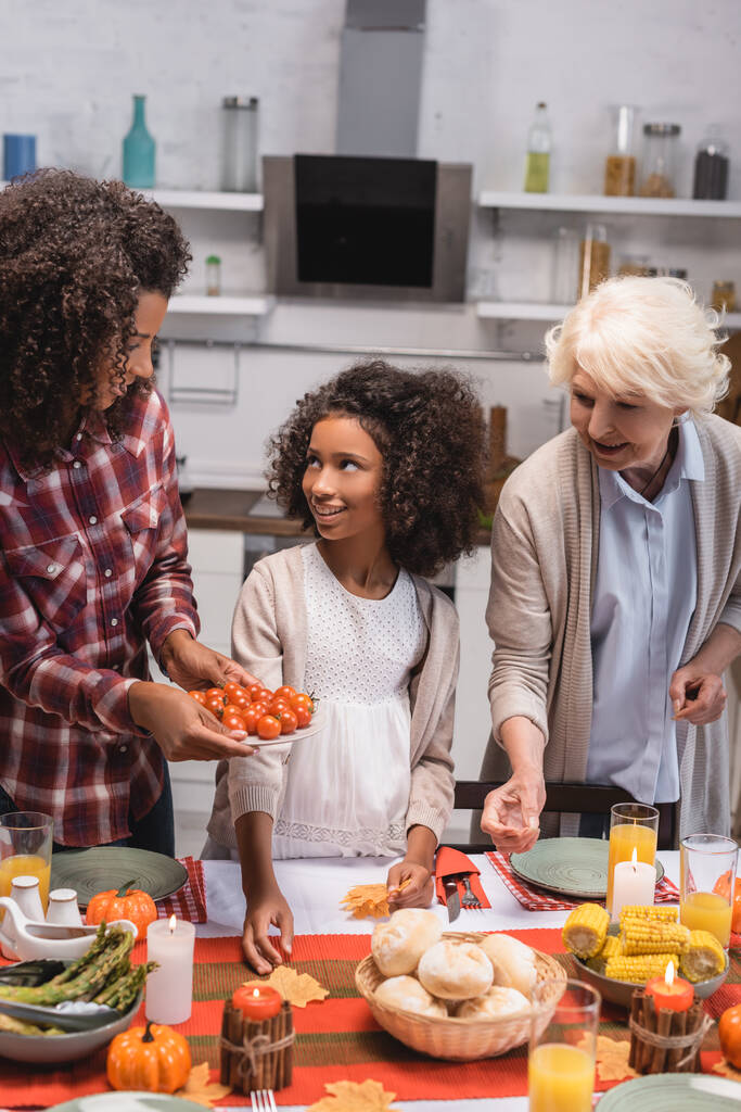 Focus selettivo della donna afro-americana che tiene i pomodorini vicino alla figlia e alla donna anziana durante il Ringraziamento  - Foto, immagini
