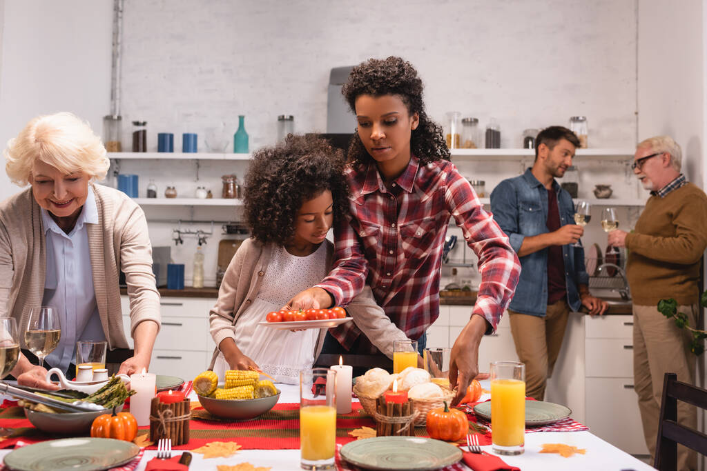 Selektiver Fokus multiethnischer Frauen, die während des Erntedankfestes Essen in der Nähe eines afrikanisch-amerikanischen Mädchens auf den Tisch bringen  - Foto, Bild