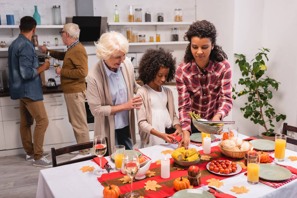 Enfoque selectivo de mujeres multiétnicas y niños de pie cerca de la comida y velas en la mesa durante el Día de Acción de Gracias  - Foto, imagen