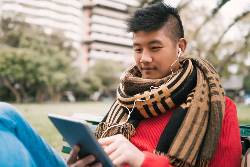 Porträt eines jungen asiatischen Mannes, der sein digitales Tablet mit Kopfhörern benutzt, während er im Freien auf einer Bank sitzt. Technologiekonzept. - Foto, Bild