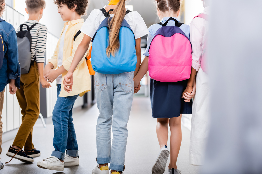 widok z tyłu wieloetnicznych kolegów trzymających się za ręce podczas spaceru szkolnym korytarzem, wybiórcze skupienie - Zdjęcie, obraz