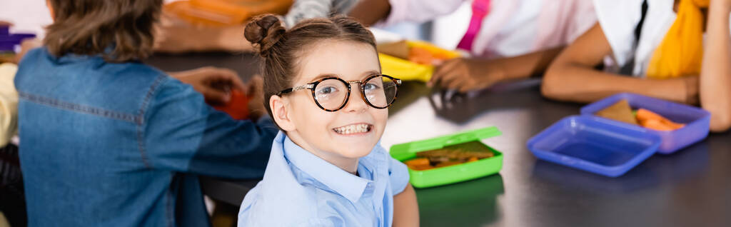 selectieve focus van opgewonden schoolmeisje in bril kijken naar camera in de buurt van klasgenoten en lunch dozen, website header - Foto, afbeelding