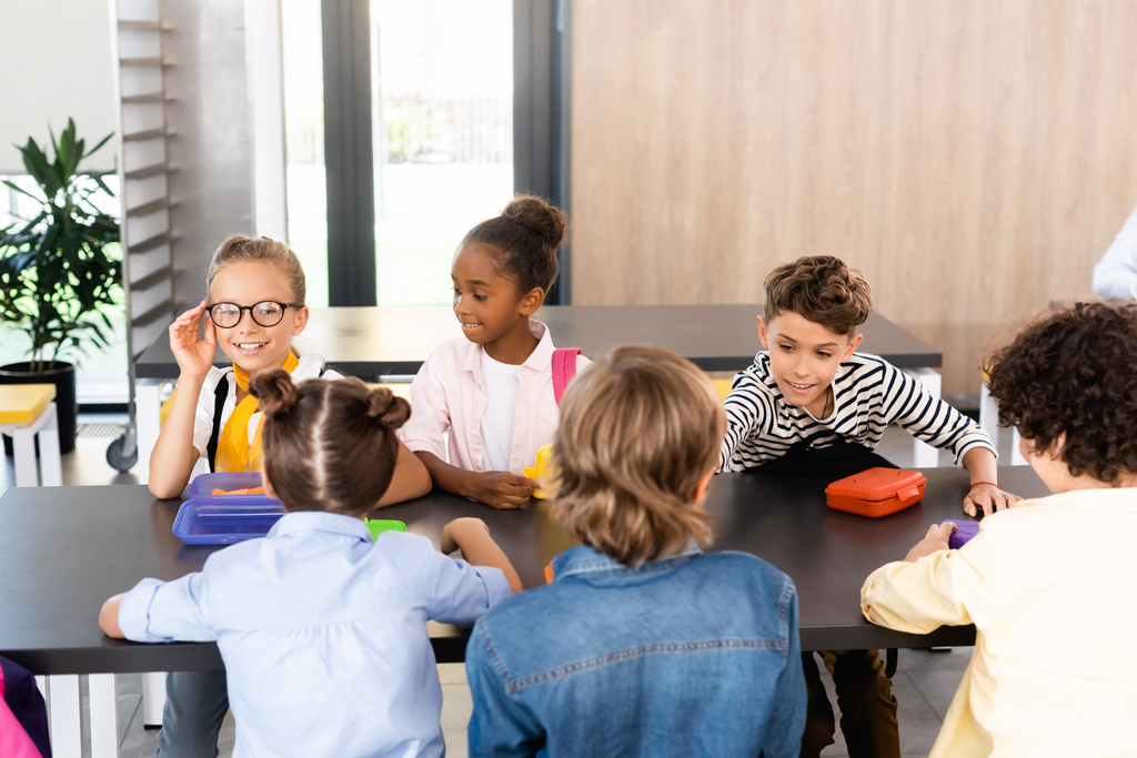colegiala tocando gafas mientras se sienta con compañeros de clase multiculturales en el comedor de la escuela - Foto, imagen