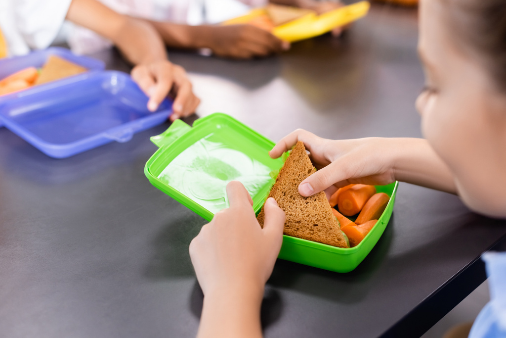Selektiver Fokus der Schülerin, die in der Nähe von Klassenkameraden Sandwich aus der Brotdose mit frischen Karotten nimmt - Foto, Bild