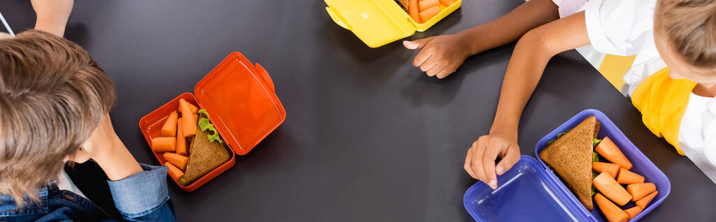 overhead view of multicultural pupils near lunch boxes with sandwiches and fresh carrots, horizontal image - Photo, Image
