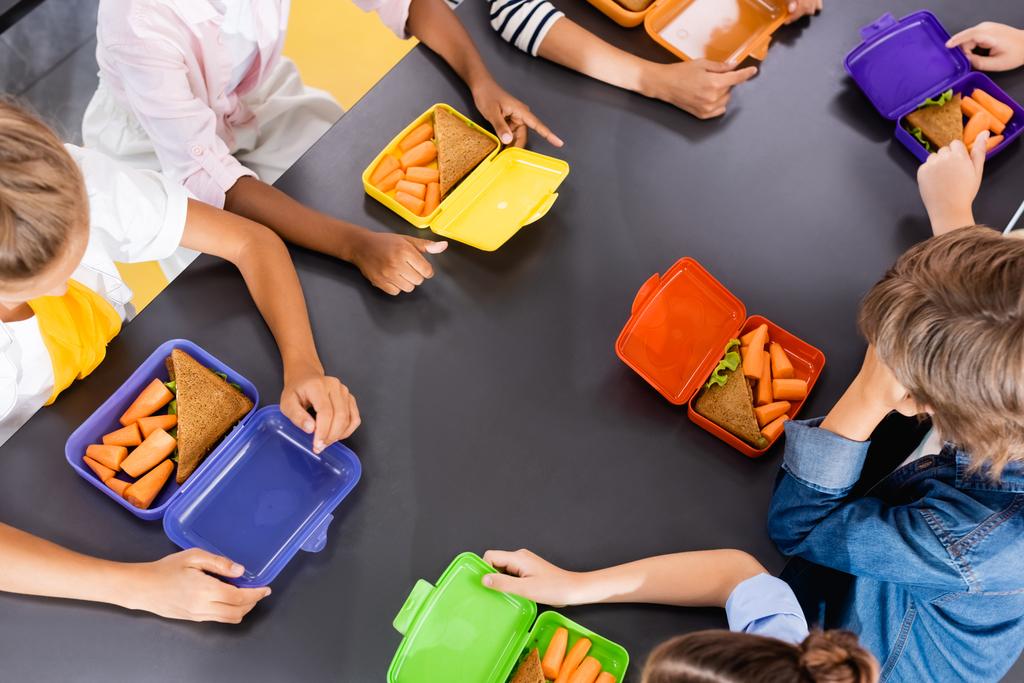 overhead view of multicultural classmates sitting in school eatery near lunch boxes with fresh carrots and sandwiches  - Photo, Image
