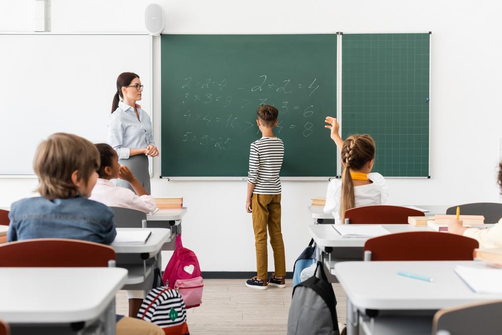 Rückseite des Schülers beim Lösen von Gleichungen auf der Tafel in der Nähe von Lehrern und multikulturellen Klassenkameraden - Foto, Bild