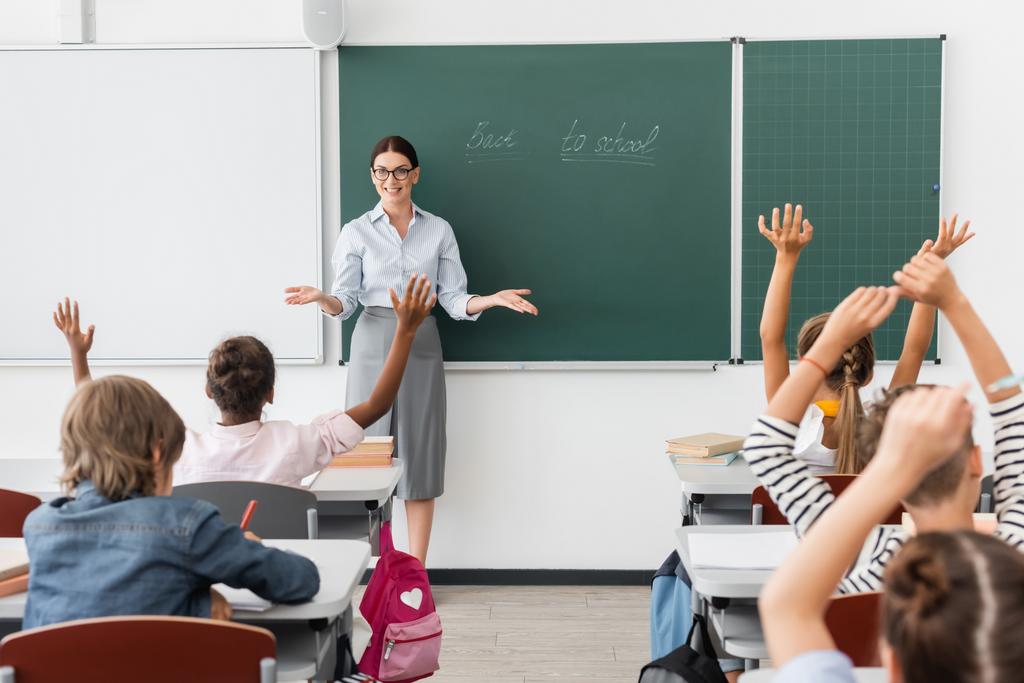 pohled zezadu na mnohonárodnostní žáky s rukama ve vzduchu a učitele stojícího s otevřenou náručí u tabule s nápisem zpět do školy - Fotografie, Obrázek