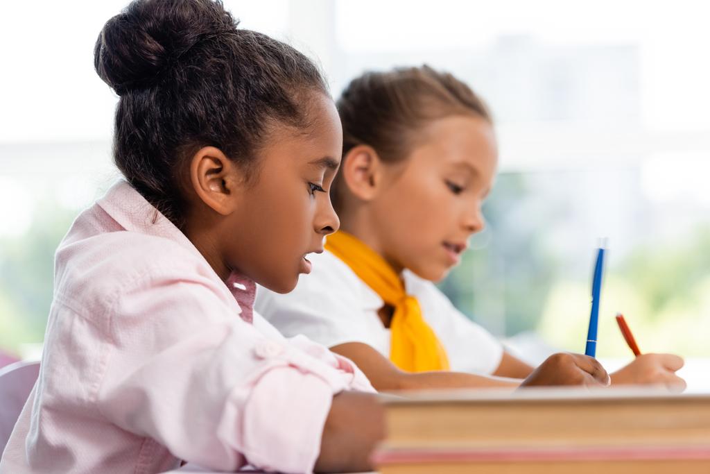 Вибіркове фокусування афроамериканської школярки, яка тримає ручку поруч з другом і книгами в класі.  - Фото, зображення
