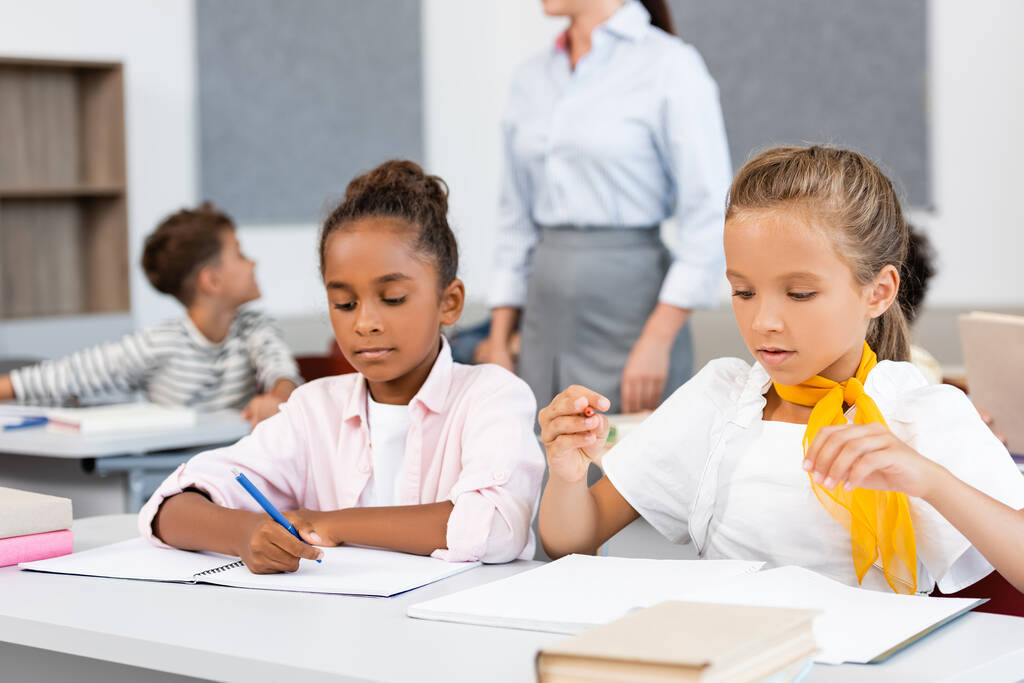 Selectieve focus van multculturele schoolmeisjes die in de buurt van boeken en notitieboekjes op een bureau in de klas zitten  - Foto, afbeelding