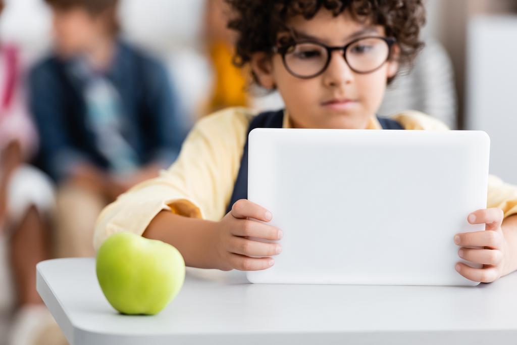 Selective focus of apple on desk near muslim schoolboy using digital tablet in school  - Photo, Image