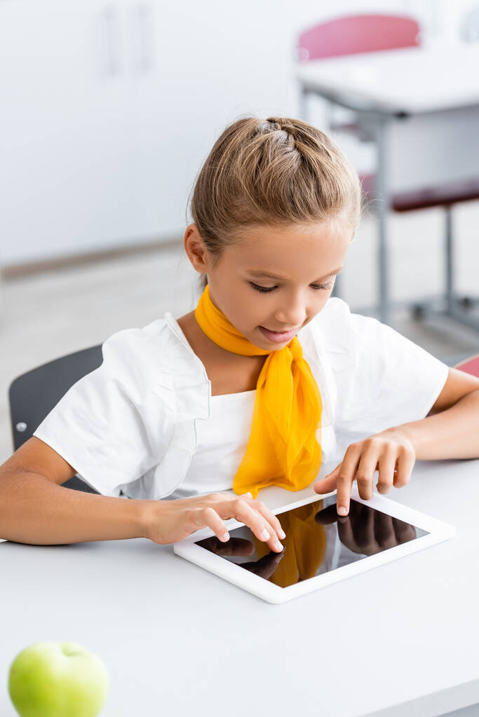 Enfoque selectivo de colegiala usando tableta digital cerca de manzana en el escritorio en el aula  - Foto, Imagen