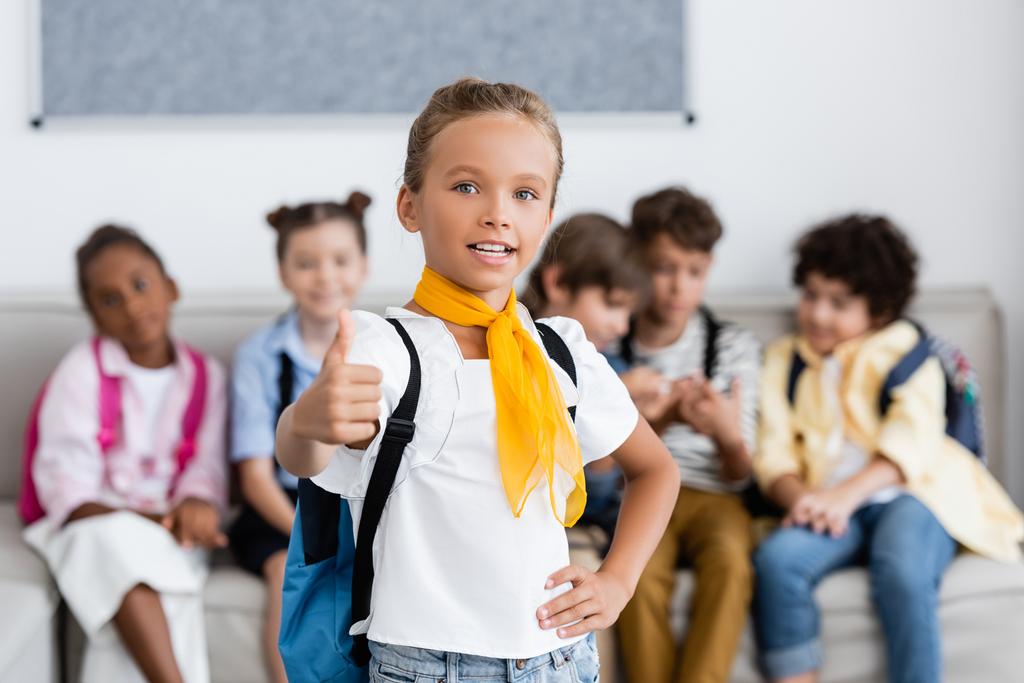 Selective focus of schoolgirl with backpack showing like gesture near multiethnic friends in school  - Photo, Image