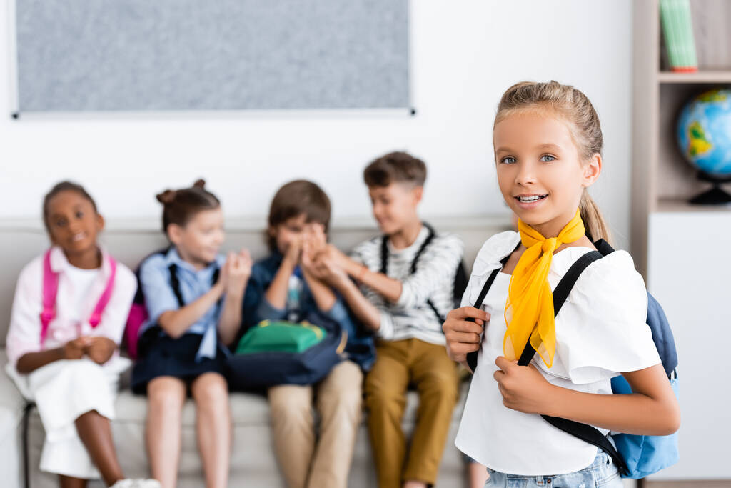 Селективный фокус школьницы с рюкзаком, смотрящей в камеру с многонациональными друзьями на заднем плане в школе  - Фото, изображение