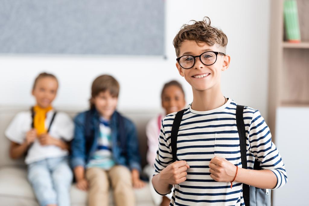 Selective focus of schoolboy with backpack and eyeglasses looking at camera with classmates at background  - Photo, Image