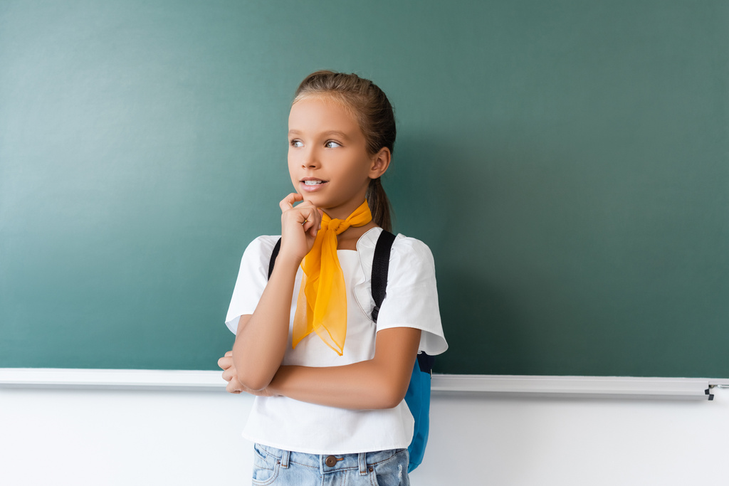Schoolgirl with backpack looking away near green chalkboard  - Photo, Image