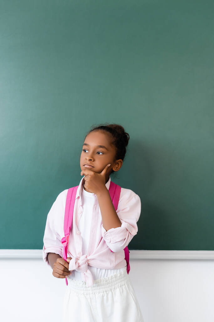 Pensive афроамериканська школярка тримає рюкзак біля зеленої дошки
  - Фото, зображення