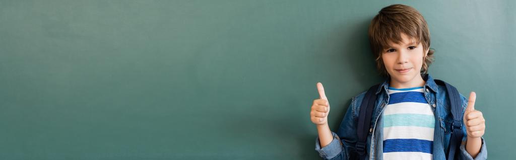 Панорамный снимок школьника, показывающий жест рядом с доской  - Фото, изображение