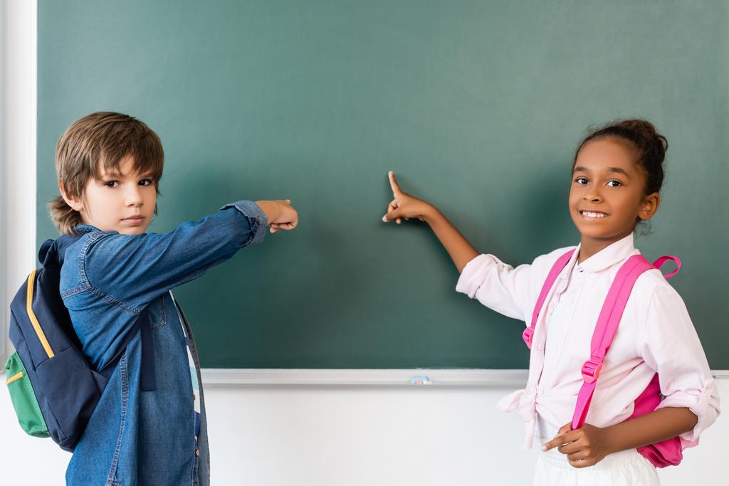 Multicultural schoolkids looking at camera while pointing at chalkboard in classroom  - Photo, Image
