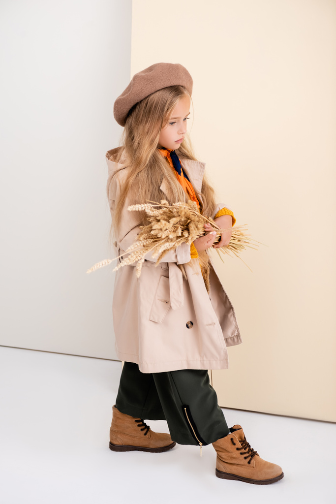 vista lateral de la chica rubia de moda en traje de otoño caminando con espigas de trigo sobre fondo beige y blanco - Foto, imagen