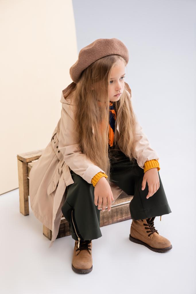 μοντέρνα ξανθιά κοπέλα σε φθινοπωρινό ντύσιμο κάθεται σε ξύλινο κουτί σε μπεζ και λευκό φόντο - Φωτογραφία, εικόνα