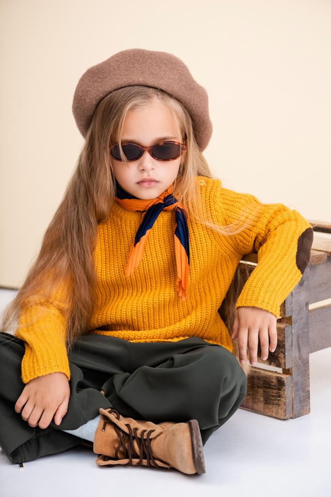 menina loira na moda em roupa de outono e óculos de sol posando perto de caixa de madeira no fundo bege e branco - Foto, Imagem