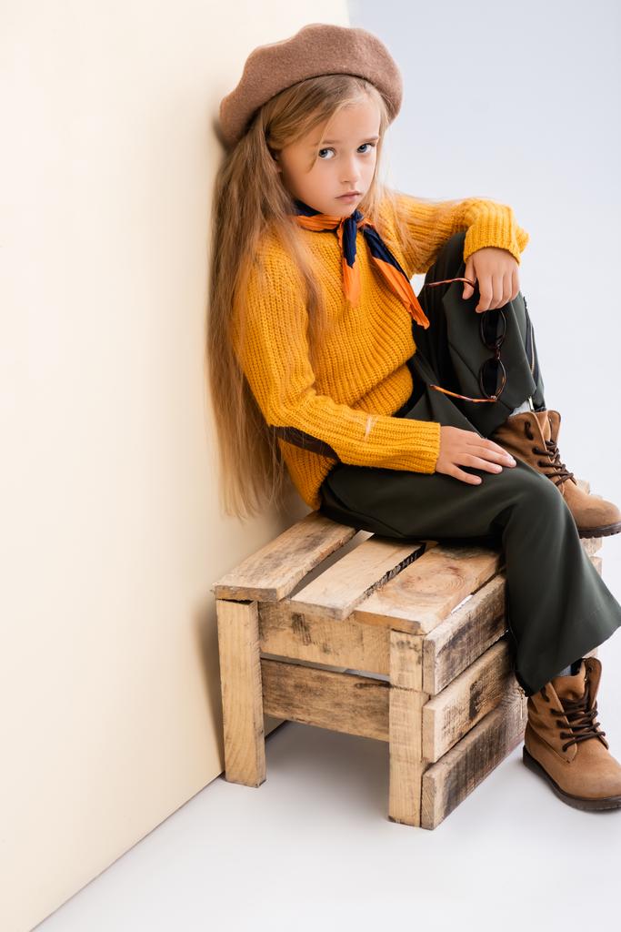 fashionable blonde girl in autumn outfit with sunglasses sitting on wooden box on beige and white background - Photo, Image