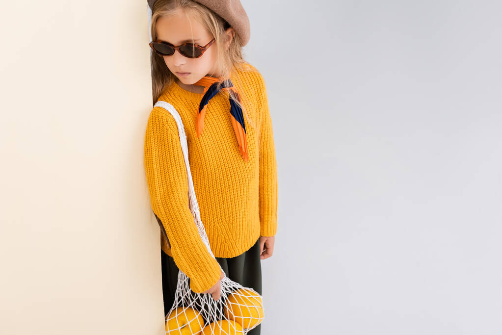 Sonbahar kıyafetli, güneş gözlüklü, bej ve beyaz arka planda greyfurtlarla poz veren şık sarışın kız. - Fotoğraf, Görsel