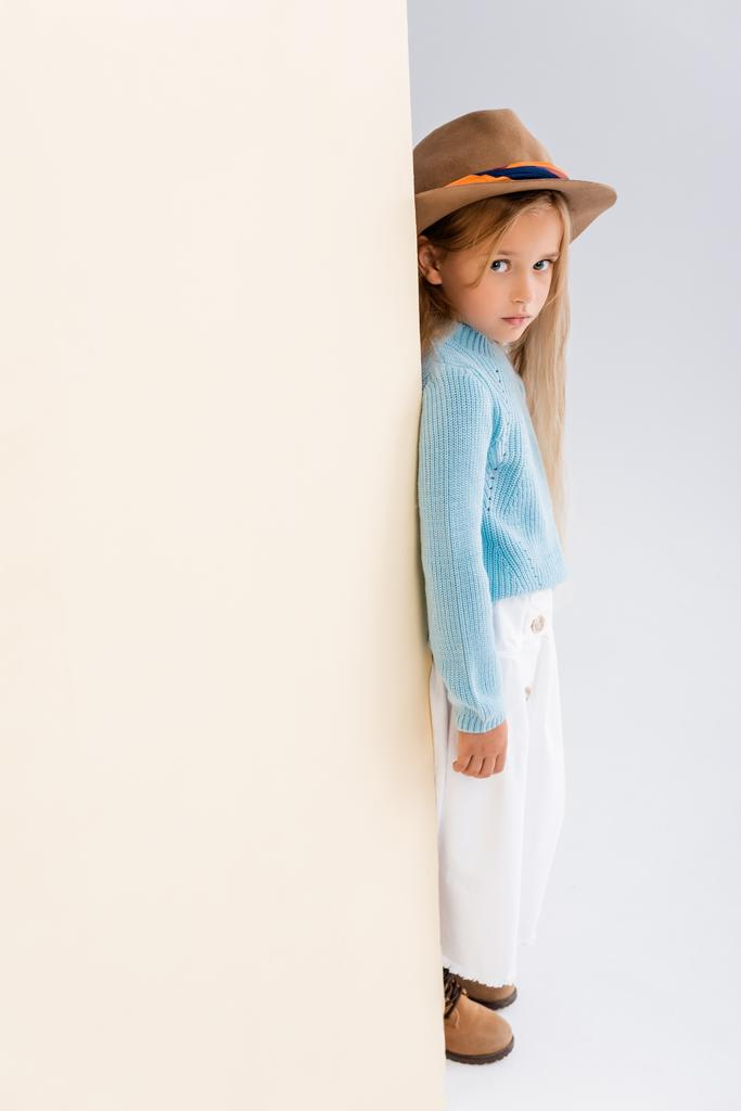 side view of fashionable blonde girl in brown hat and boots, white skirt and blue sweater near beige wall - Photo, Image