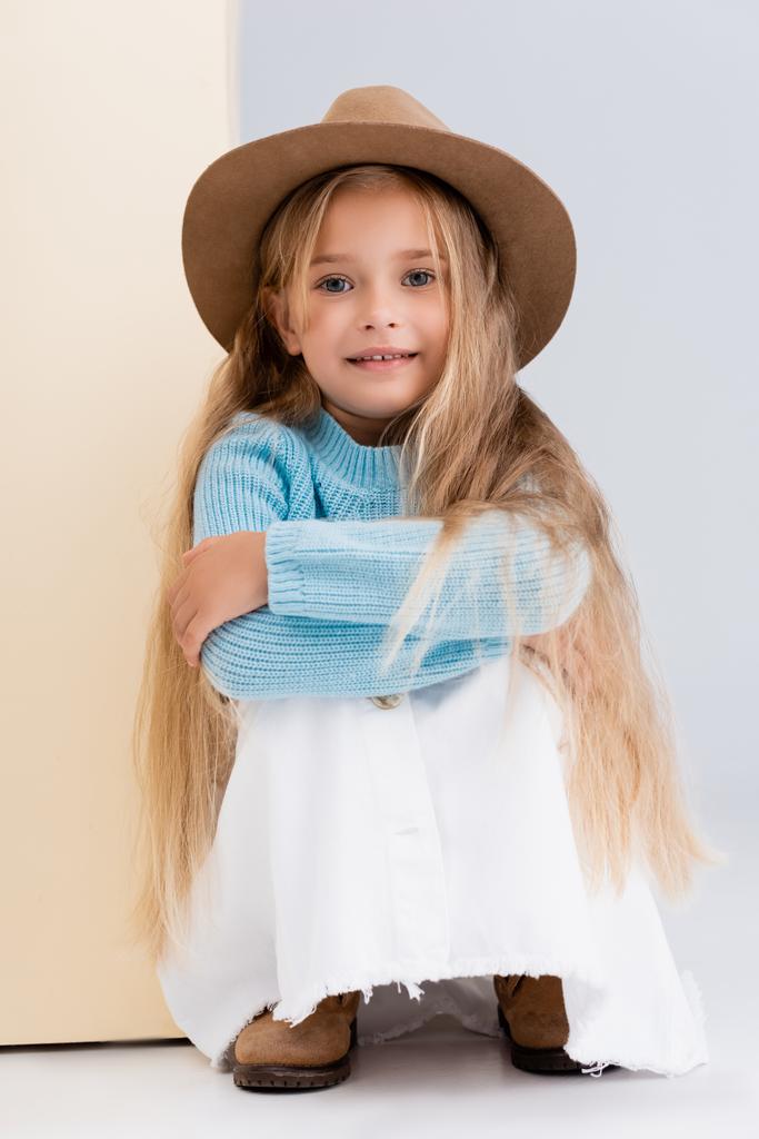 modisches blondes Mädchen mit braunem Hut und Stiefeln, weißem Rock und blauem Pullover in der Nähe der beigen Wand - Foto, Bild