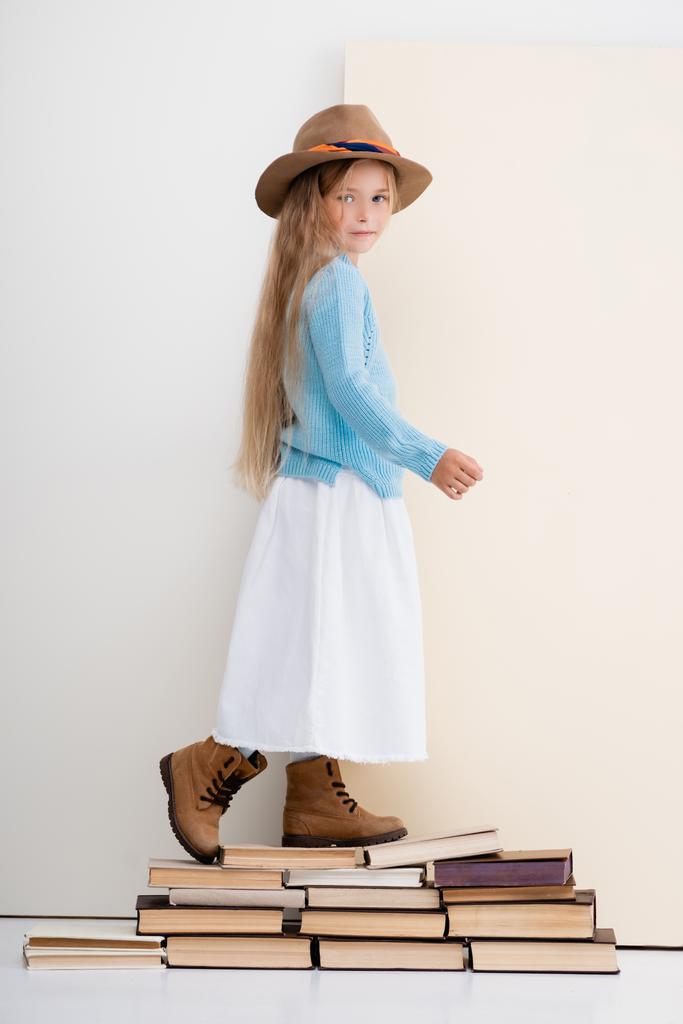 Seitenansicht des modischen blonden Mädchens mit braunem Hut und Stiefeln, weißem Rock und blauem Pullover, das auf alten Büchern wandelt - Foto, Bild