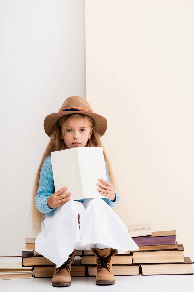 modna blondynka dziewczyna w brązowym kapeluszu i buty, biała spódnica i niebieski sweter siedzi z książki w pobliżu beżowej ściany - Zdjęcie, obraz