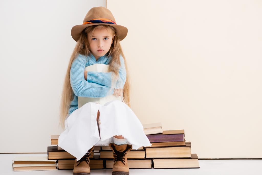 triste fille blonde à la mode en chapeau brun et bottes, jupe blanche et pull bleu assis avec livre près du mur beige - Photo, image