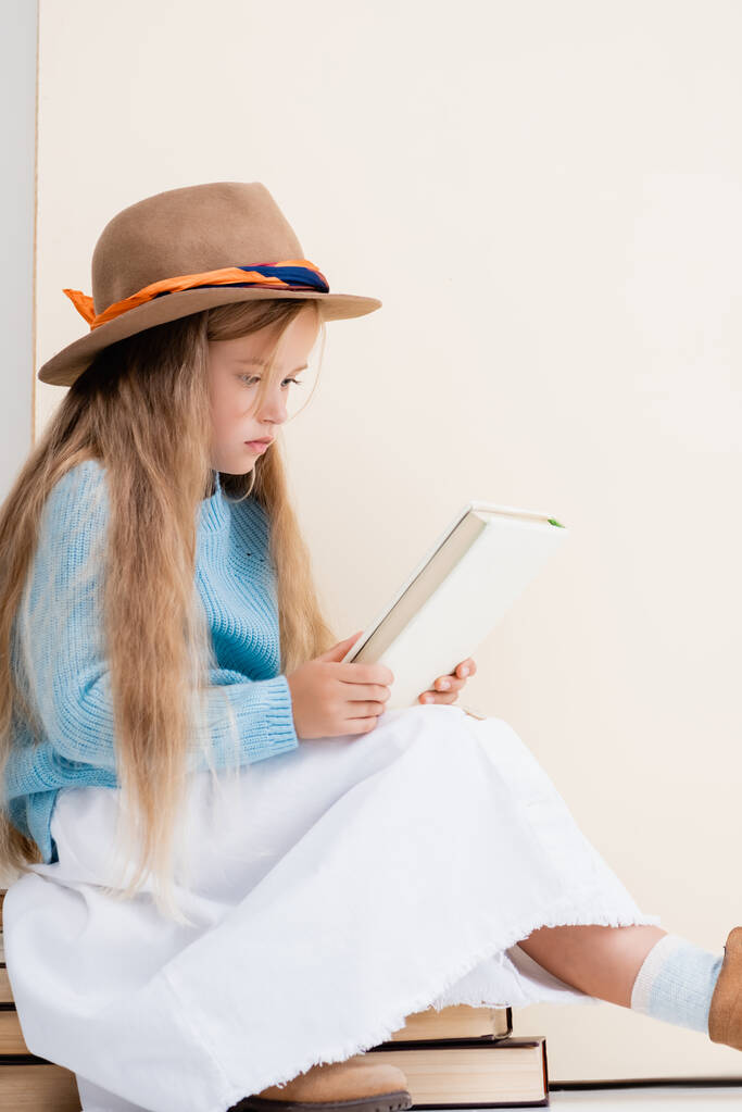 fashionable blonde girl in brown hat, white skirt and blue sweater sitting on vintage books and reading near beige wall - Photo, Image