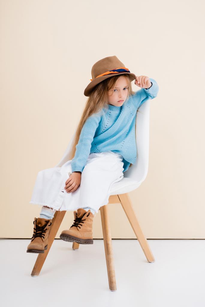 модная блондинка в коричневой шляпе и сапогах, белая юбка и голубой свитер сидят на стуле возле бежевой стены - Фото, изображение