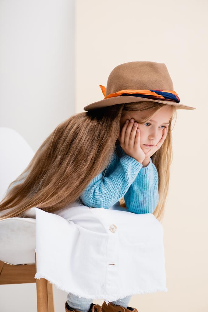 sad fashionable blonde girl in brown hat, white skirt and blue sweater sitting on chair near beige wall - Photo, Image