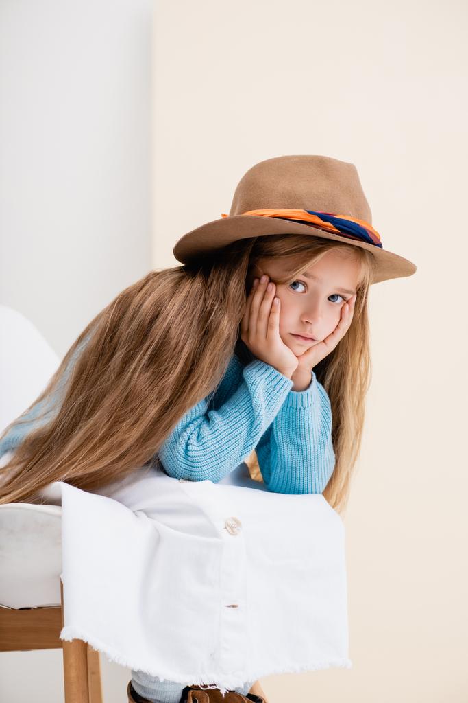 sad fashionable blonde girl in brown hat, white skirt and blue sweater sitting on chair near beige wall - Photo, Image