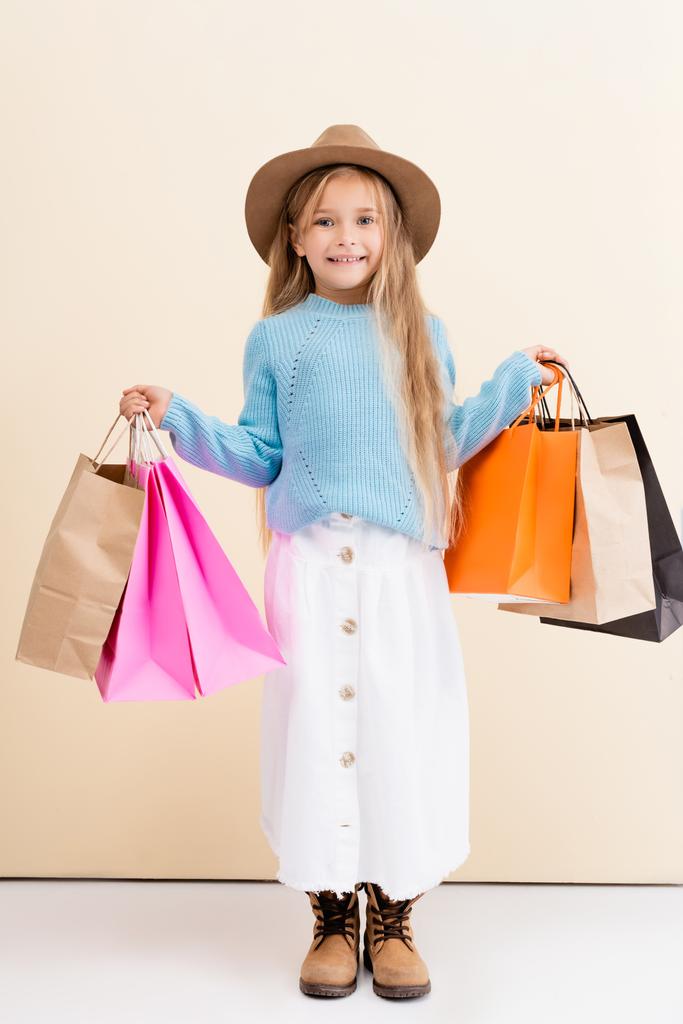 módní blondýnka v hnědém klobouku a boty, bílá sukně a modrý svetr s barevnými nákupními taškami s úsměvem v blízkosti béžové stěny - Fotografie, Obrázek