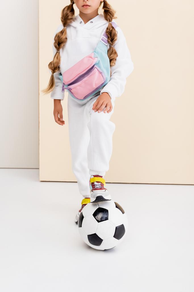 vista ritagliata di ragazza bionda in abbigliamento sportivo in posa con pallone da calcio su sfondo beige e bianco - Foto, immagini