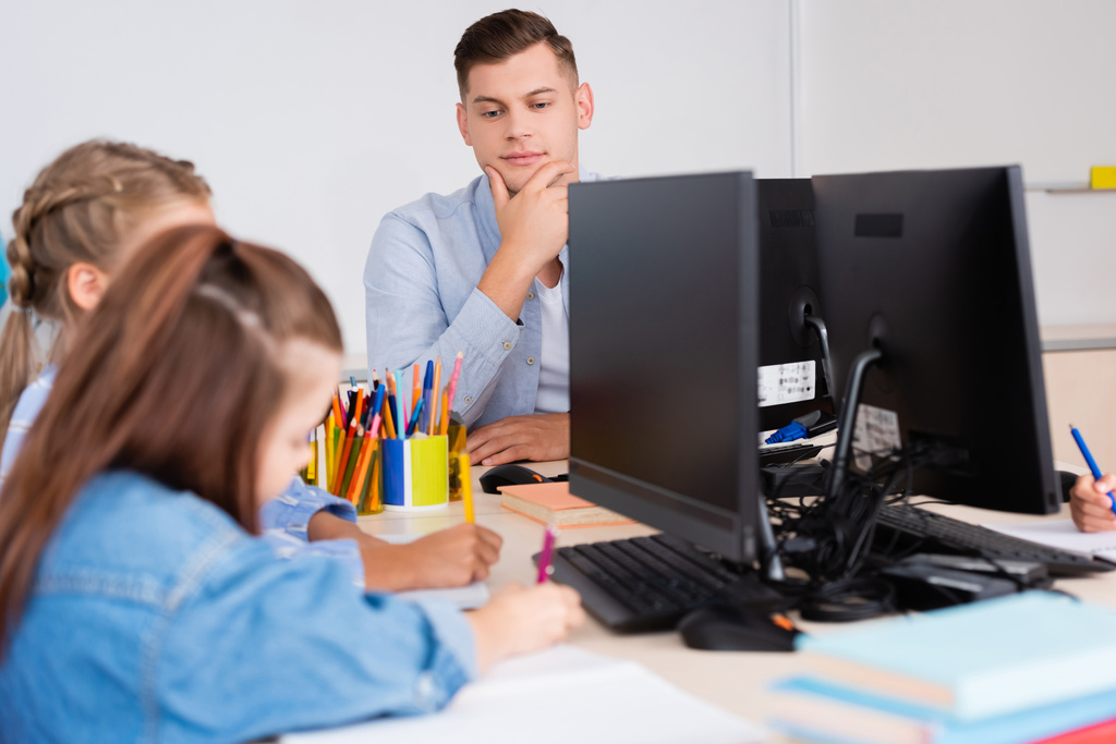 Επιλεκτική εστίαση του δασκάλου που κάθεται κοντά σε μαθητές γράφοντας σε σημειωματάρια κοντά σε υπολογιστές στο σχολείο  - Φωτογραφία, εικόνα
