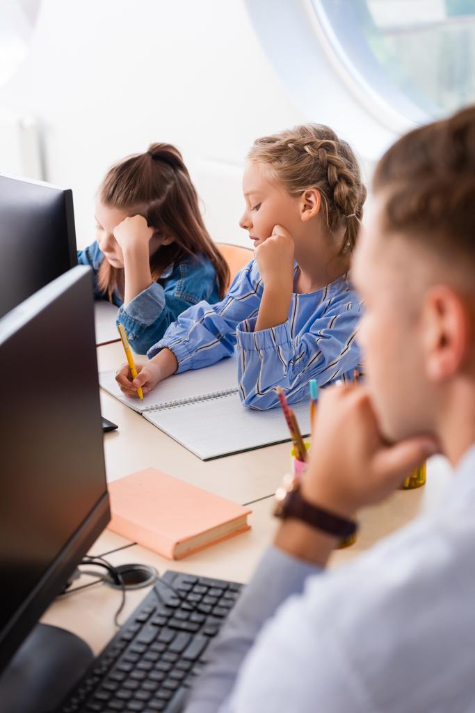 Szelektív fókusz iskoláslányok írás notebook közelében tanár és számítógépek osztályteremben  - Fotó, kép