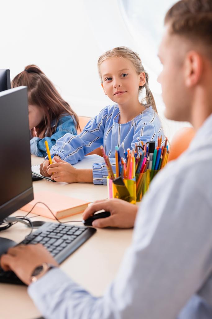 Focus selettivo della studentessa con penna che guarda l'insegnante vicino ai computer in classe  - Foto, immagini