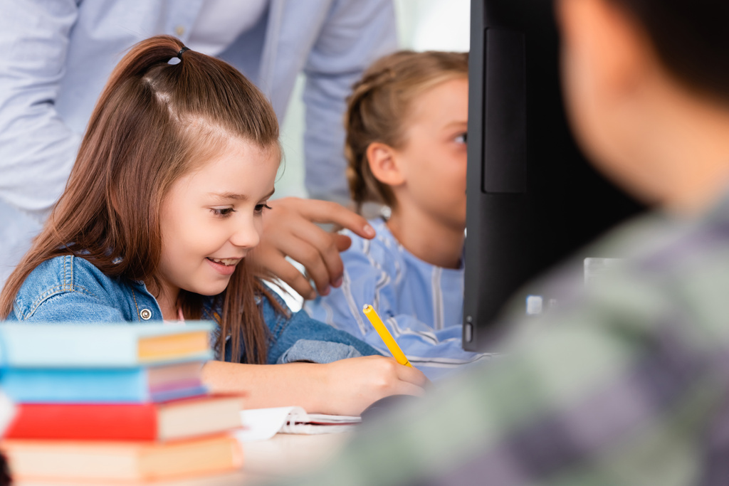 Селективный фокус учителя, стоящего рядом с школьницей, пишущей на блокноте в начальной школе  - Фото, изображение