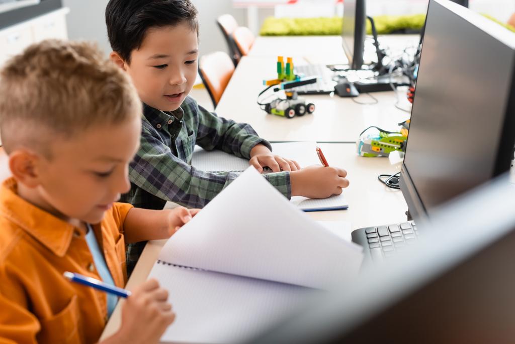 Селективный фокус мультиэтнических школьников с ноутбуками, сидящими рядом с компьютерами в начальной школе  - Фото, изображение