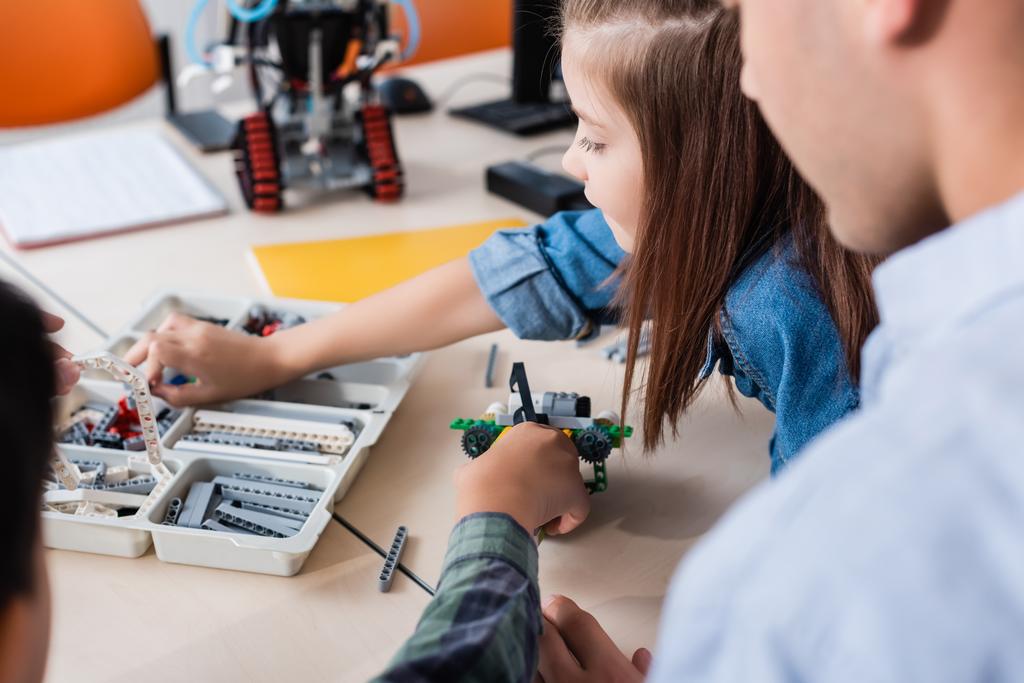 Selective focus of teacher sitting near schoolkids modeling robot in classroom  - Photo, Image