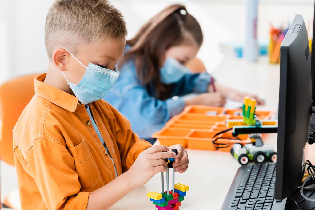 Szelektív fókusz iskolás fiú orvosi maszk modellezés robot építőkockák és számítógép az iskolában  - Fotó, kép