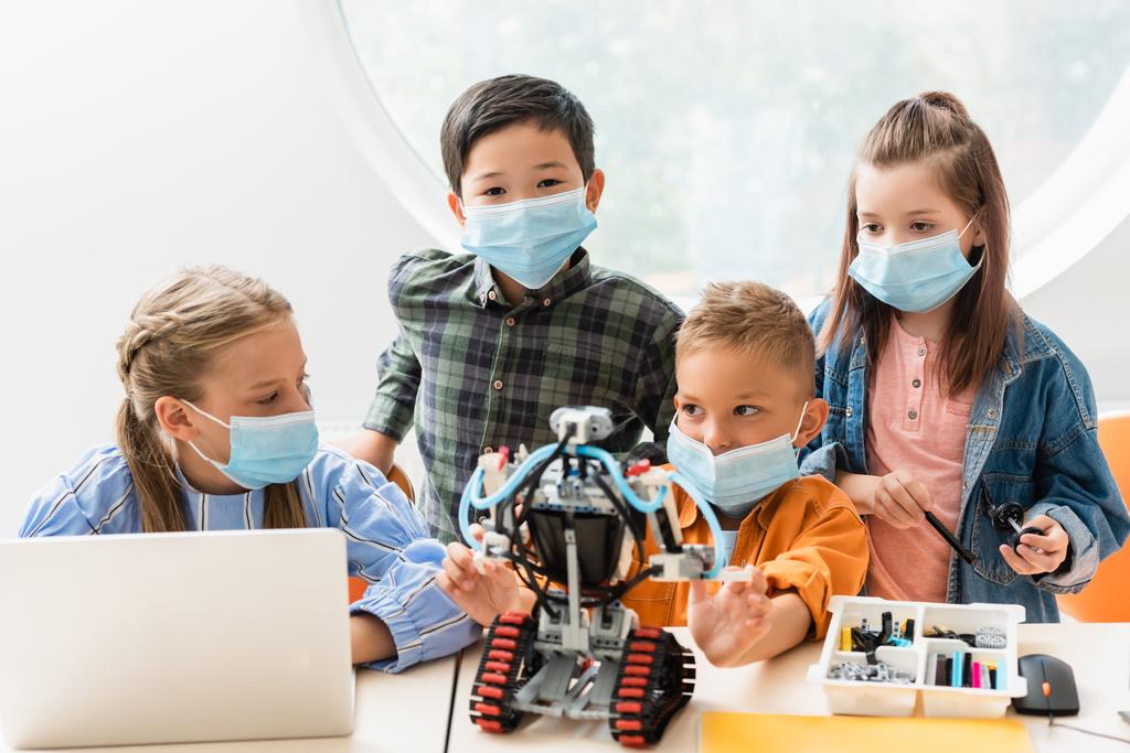 Вибірковий фокус багатоетнічних однокласників у медичних масках конструювання робота поблизу ноутбука в класі
  - Фото, зображення