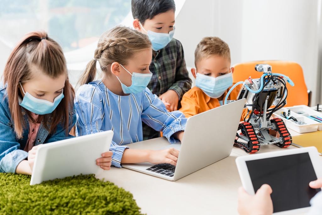Επιλεκτική εστίαση πολυεθνικών παιδιών με ιατρικές μάσκες με τη χρήση ψηφιακών tablet και laptop κοντά στο δάσκαλο και το ρομπότ στο σχολείο  - Φωτογραφία, εικόνα