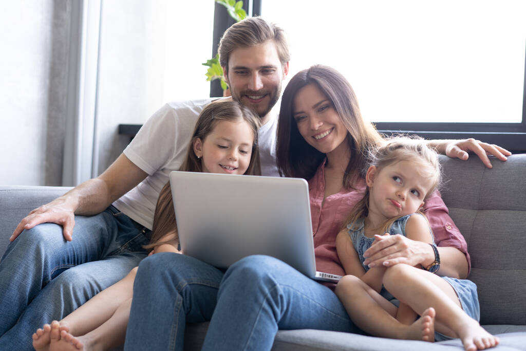 笑顔の両親と子供たちは自宅でソファでノートパソコンを使用して、コンピュータで面白いビデオを楽しんで - 写真・画像